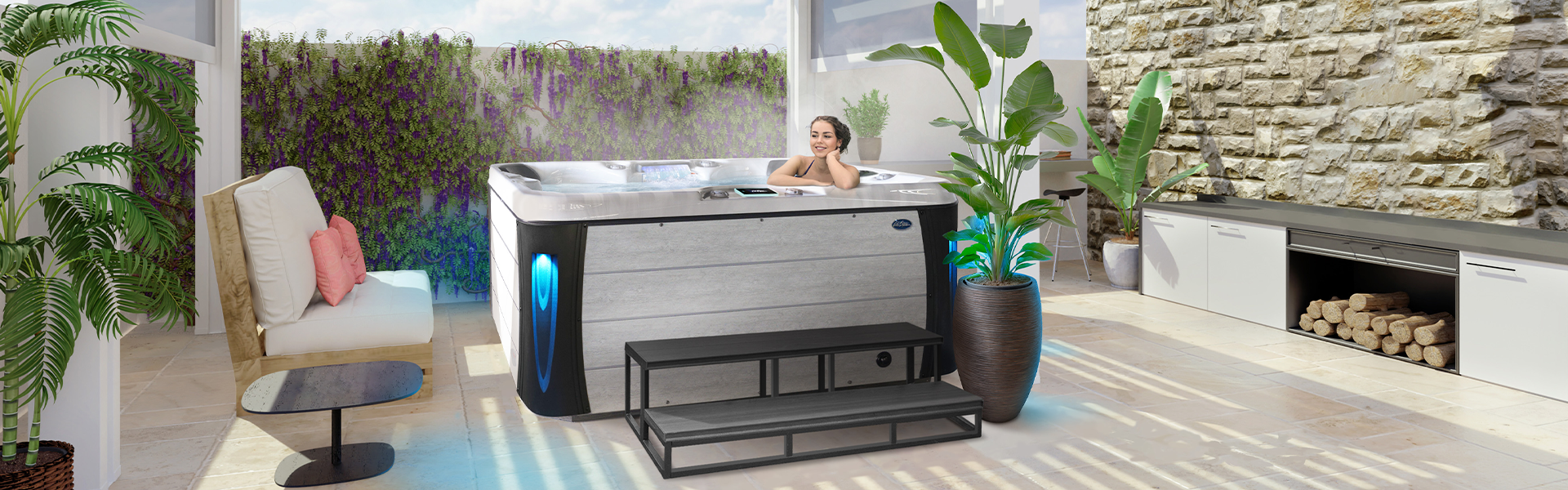 A person lounging in a Cal Spas Excape X hot tub with lime oak cabinet, blue lighted corners and 60-inch steps, in an outdoor living space.